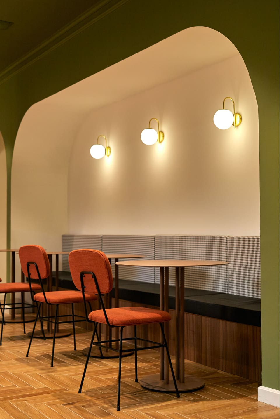 Sitzbereich mit grüner Wand, gerundeter Öffnung, Wandleuchten und orangefarbenen Stühlen.